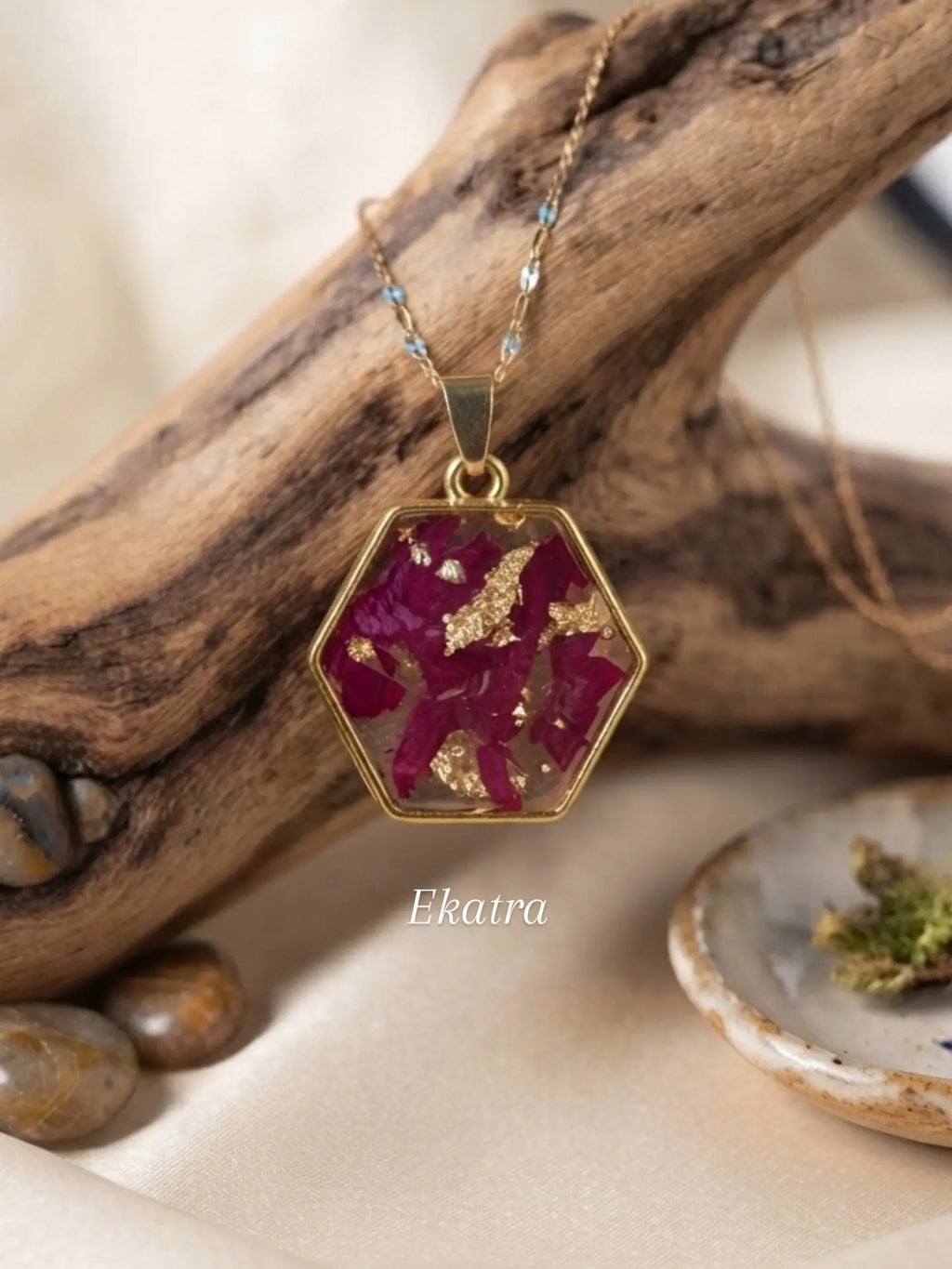 A gold hexagonal pendant necklace with dried magenta-pink flowers and gold leaf, displayed on a piece of driftwood with a small dish of dried herbs. The brand name 'Ekatra' is visible as a watermark.