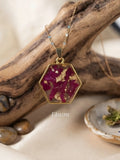 A gold hexagonal pendant necklace with dried magenta-pink flowers and gold leaf, displayed on a piece of driftwood with a small dish of dried herbs. The brand name 'Ekatra' is visible as a watermark.