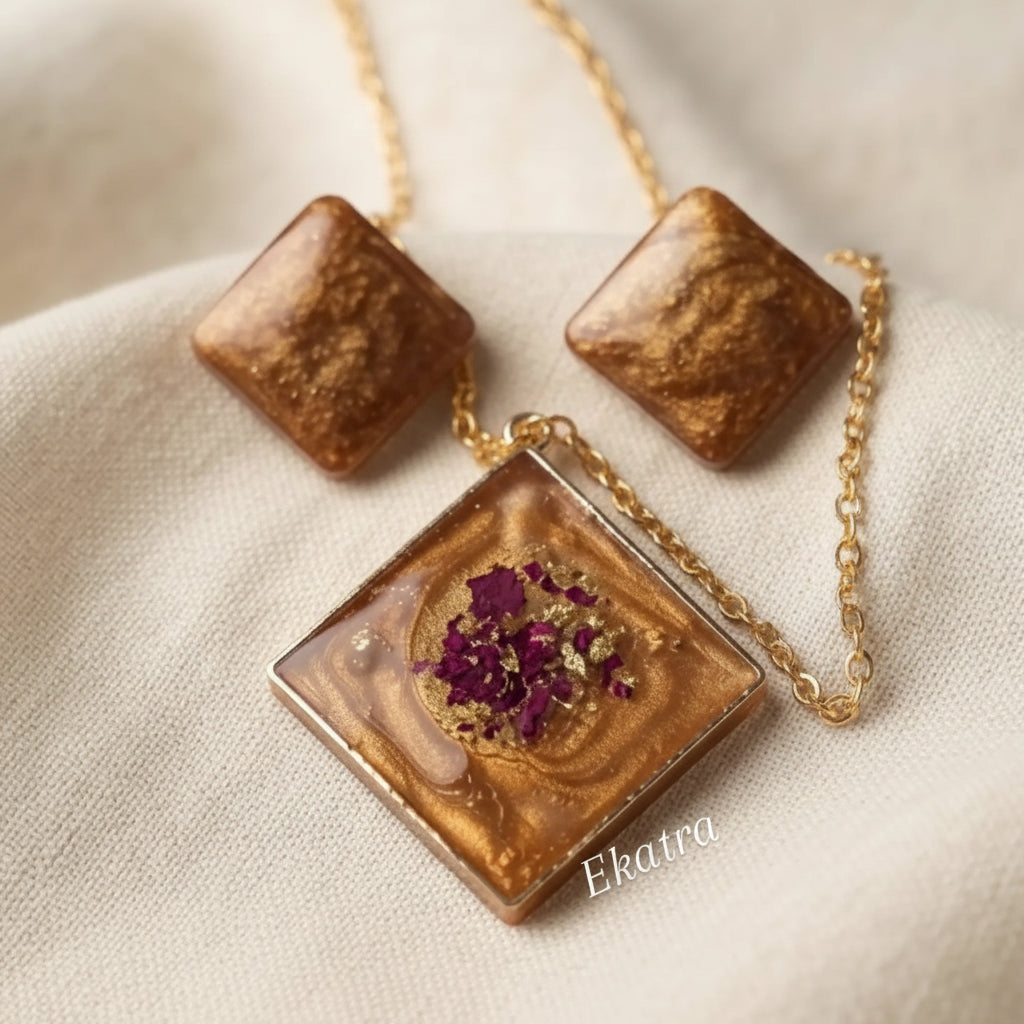 A jewelry set featuring a square pendant and matching square stud earrings with a marbled amber-brown resin design, gold foil flakes, and a dried purple flower in the center, displayed on a textured beige fabric background.