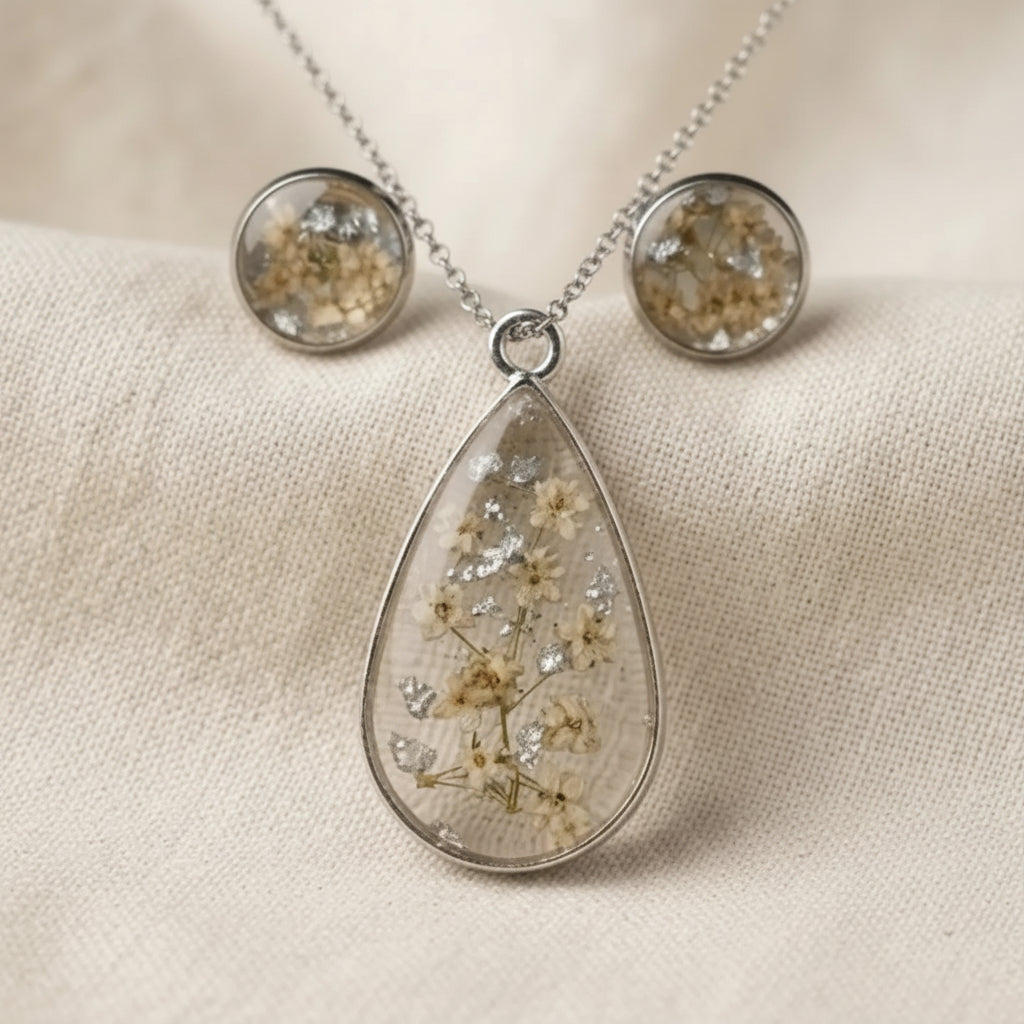 A teardrop-shaped pendant necklace and matching round stud earrings with embedded dried flowers and glitter in a clear resin, displayed on a beige fabric background. The pendant has a silver-tone border and bail, and the studs have matching silver-tone settings.