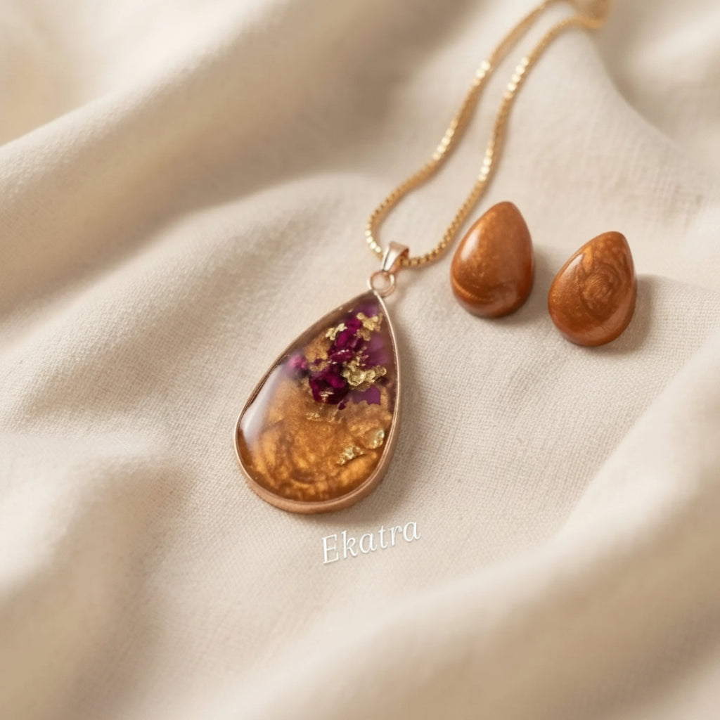 A teardrop-shaped pendant with a warm amber-brown marbled pattern, purple dried flower inclusions, and gold foil flakes, displayed next to two matching brown beads on a draped cream fabric background. The brand name 'Ekatra' is printed in white text near the bottom.