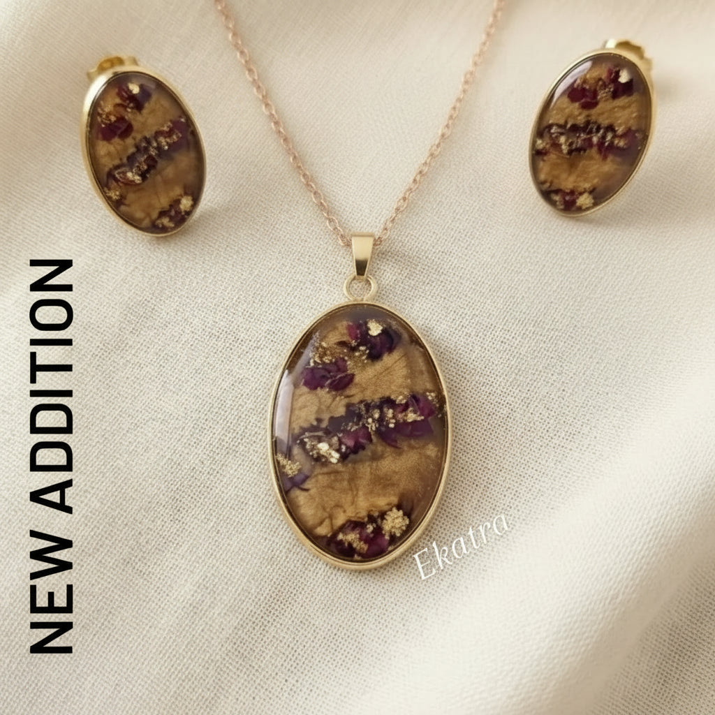 A jewelry set featuring an oval pendant and matching stud earrings with a brown and purple dried floral resin inlay, set in gold-tone metal on a beige fabric background.